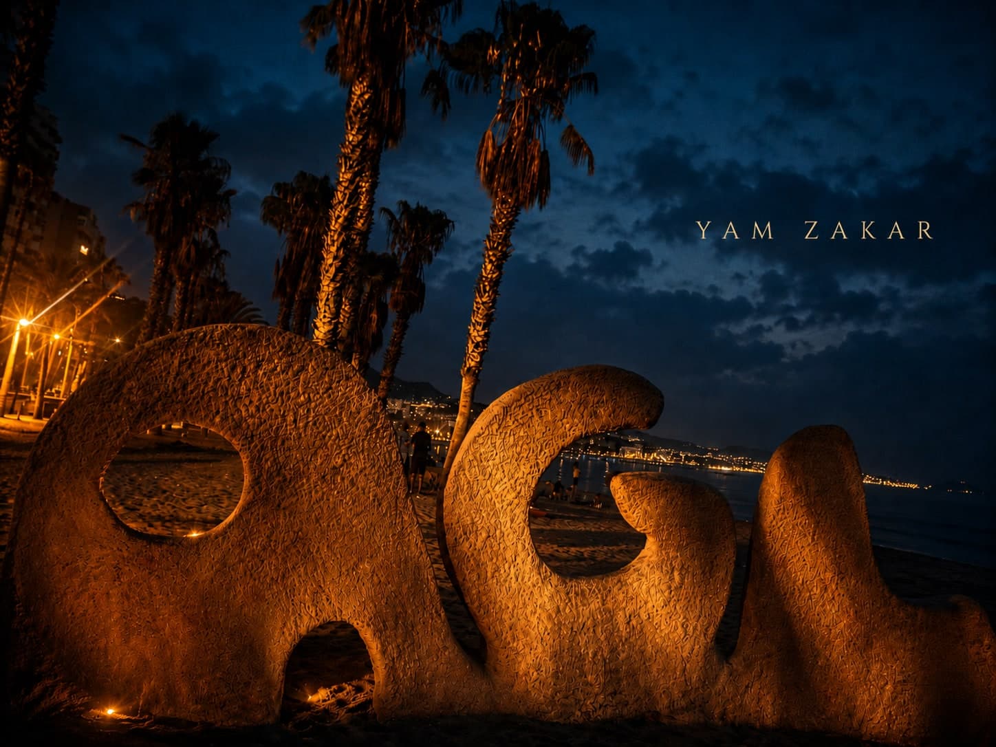A close crop of the word MALAGUETA sculpted in sand on the beach at Málaga, lit gold from below at night. Only the middle letters are in frame. Palms and a curve of city lights stand behind them, and the words YAM ZAKAR are set in the dark blue sky.